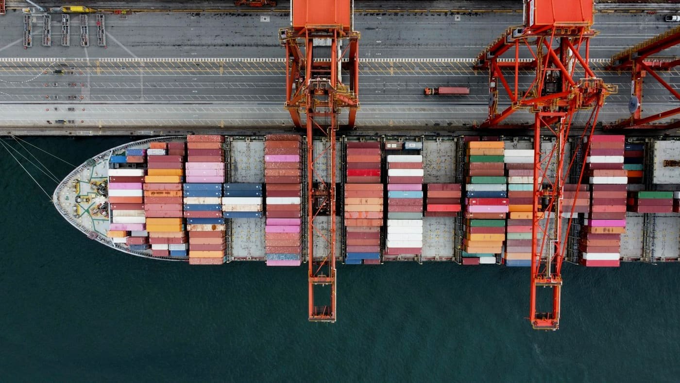 Aerial view of a container ship loaded with colourful containers beside two quay cranes, illustrating sea freight operations