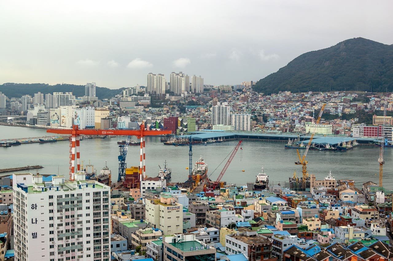Harbour with port cranes below a hillside city, labelled Busan by the photographer, illustrating South Korea to Russia shipping from Busan