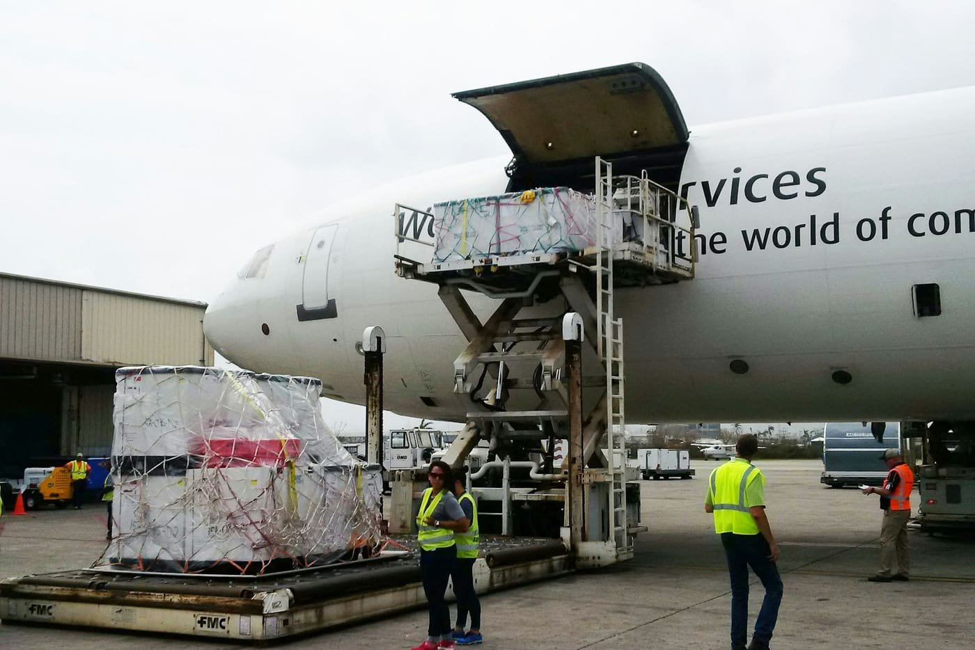 Cargo aircraft with its nose door open as a pallet of freight is raised on a loader beside ground crew in yellow vests