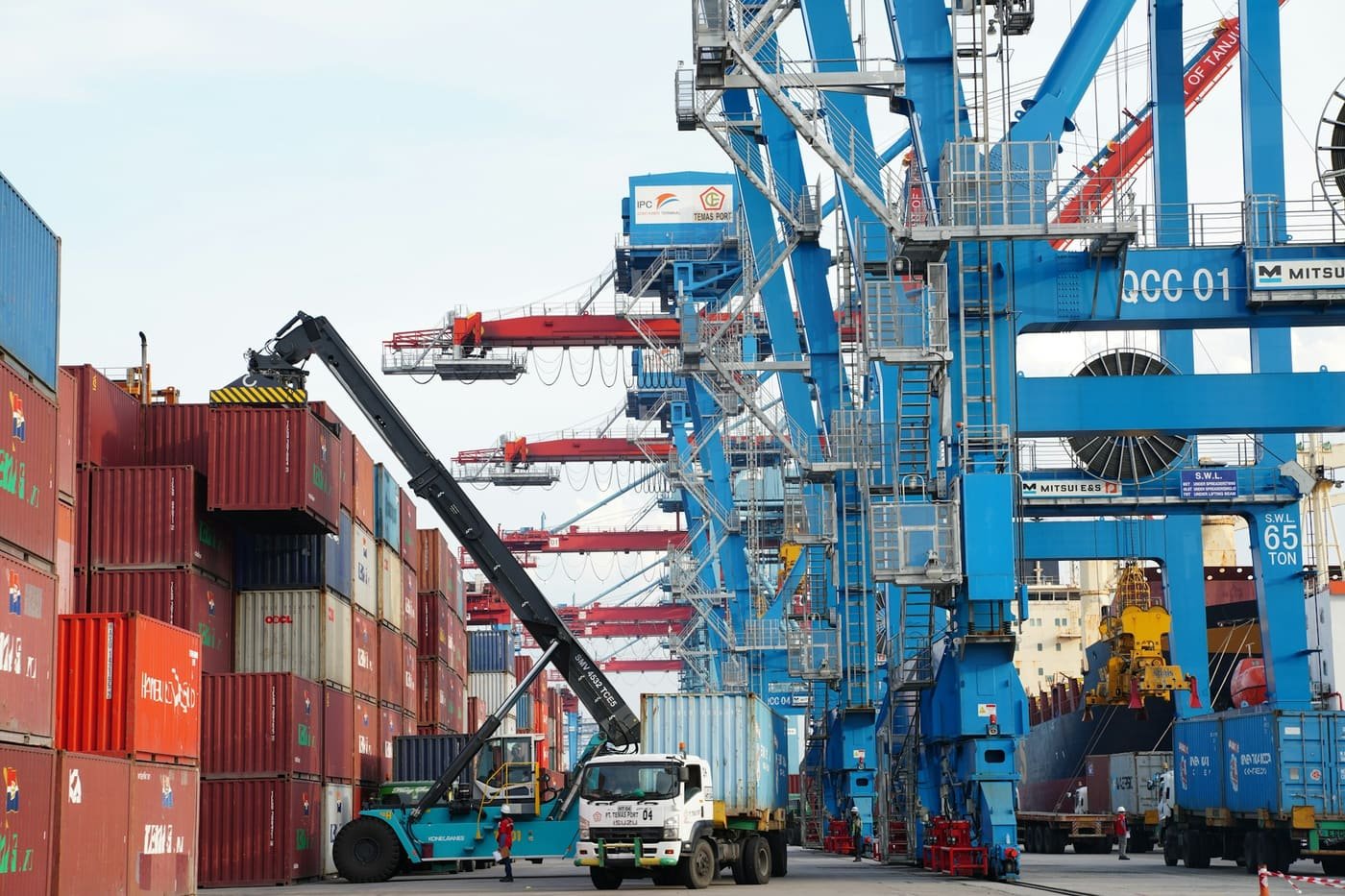 Container handler and truck working among container stacks beneath blue quay cranes