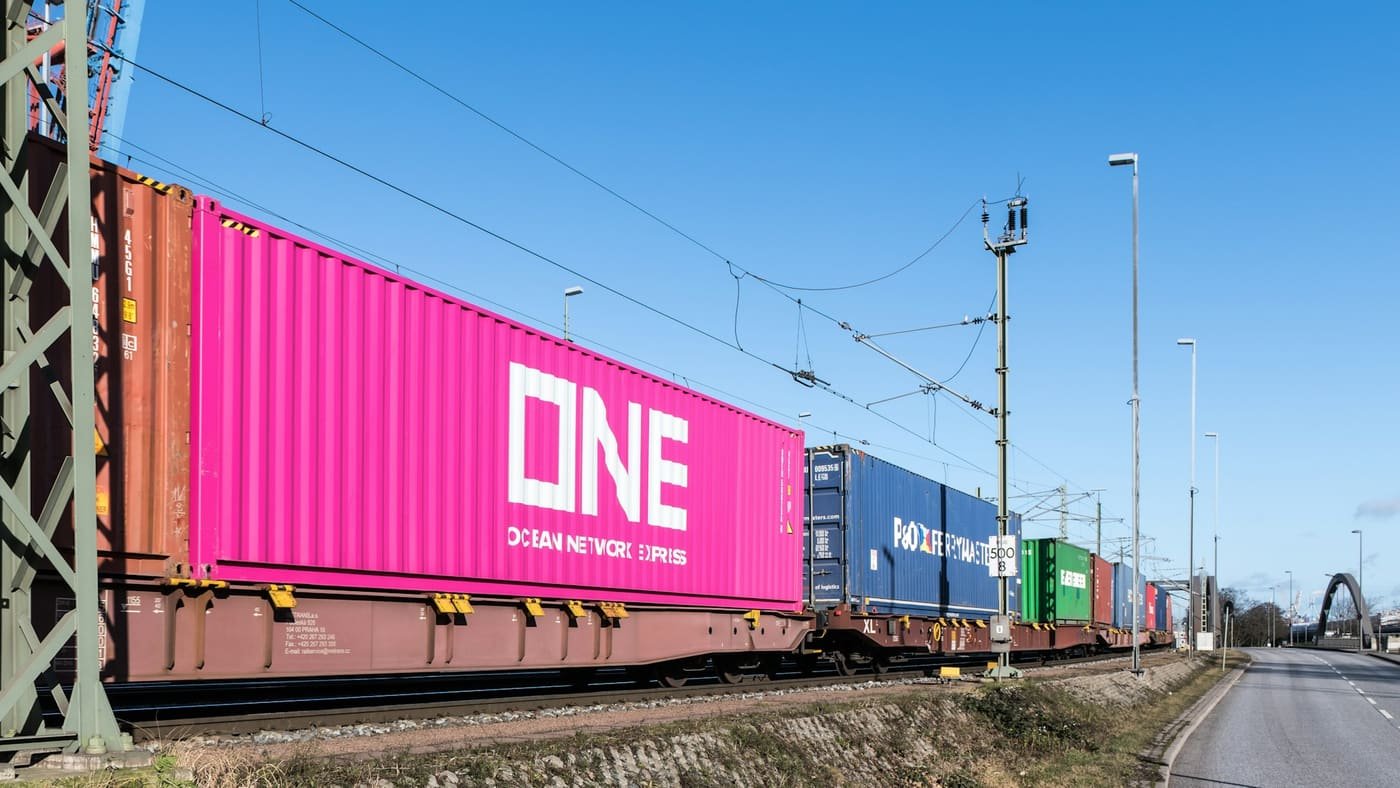 Freight train carrying pink, blue and green shipping containers on rail wagons beside a road