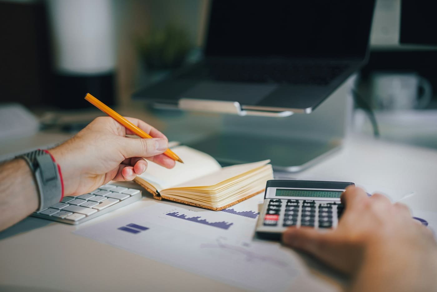 Hands using a calculator and writing in a notebook beside a laptop