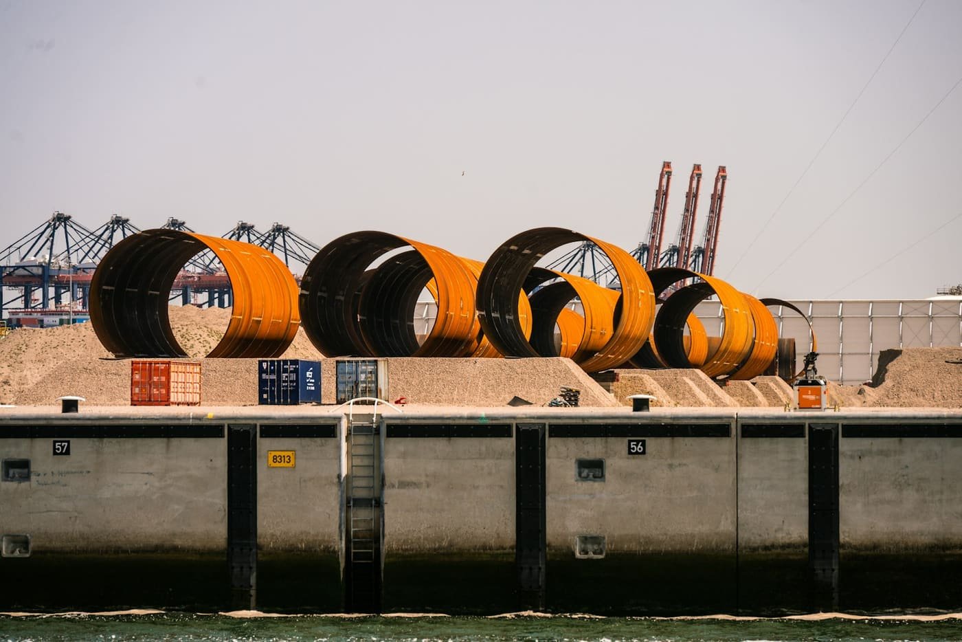 Large orange steel pipe sections on a quay with port cranes behind