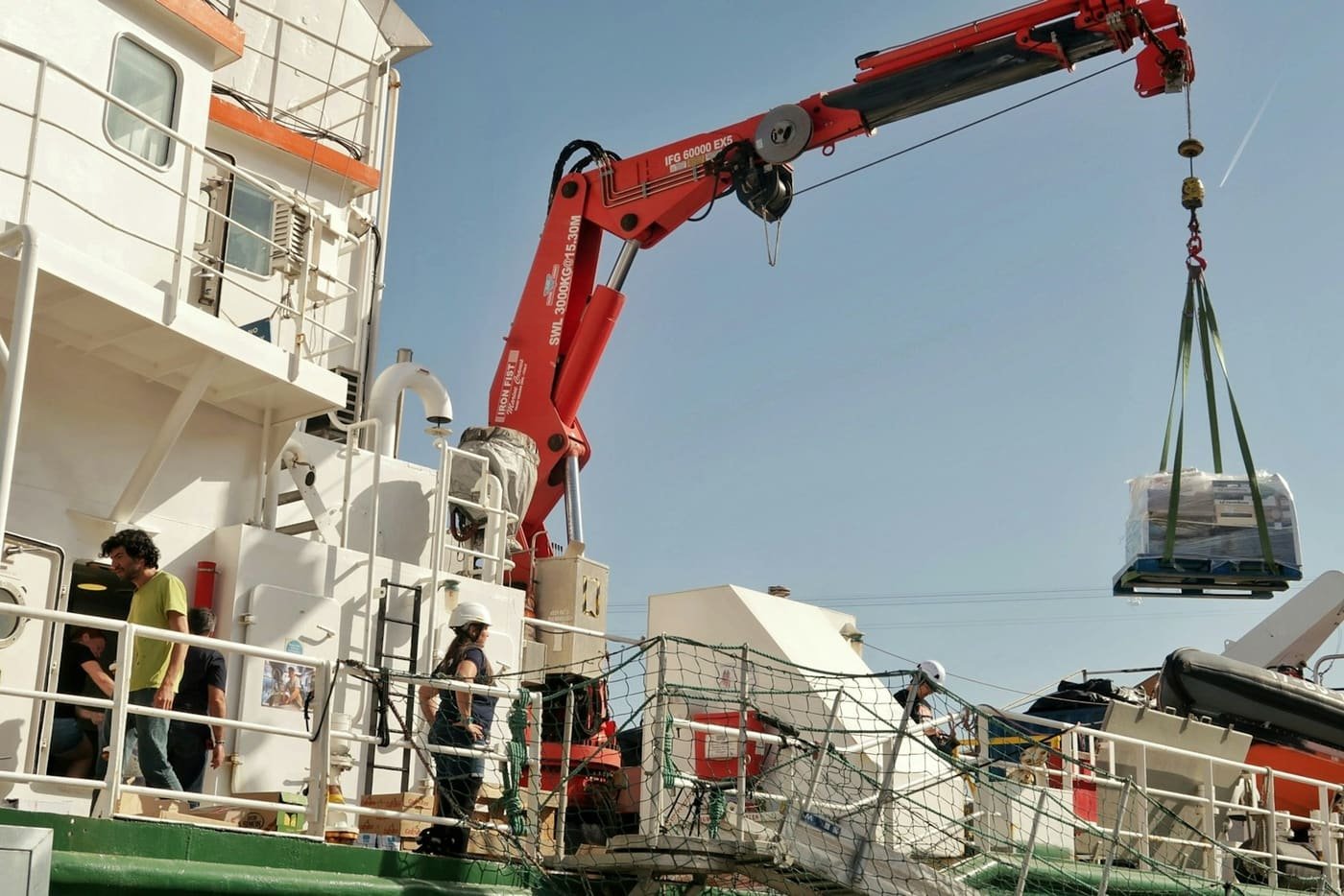 Ship's crane lifting a wrapped pallet while crew look on