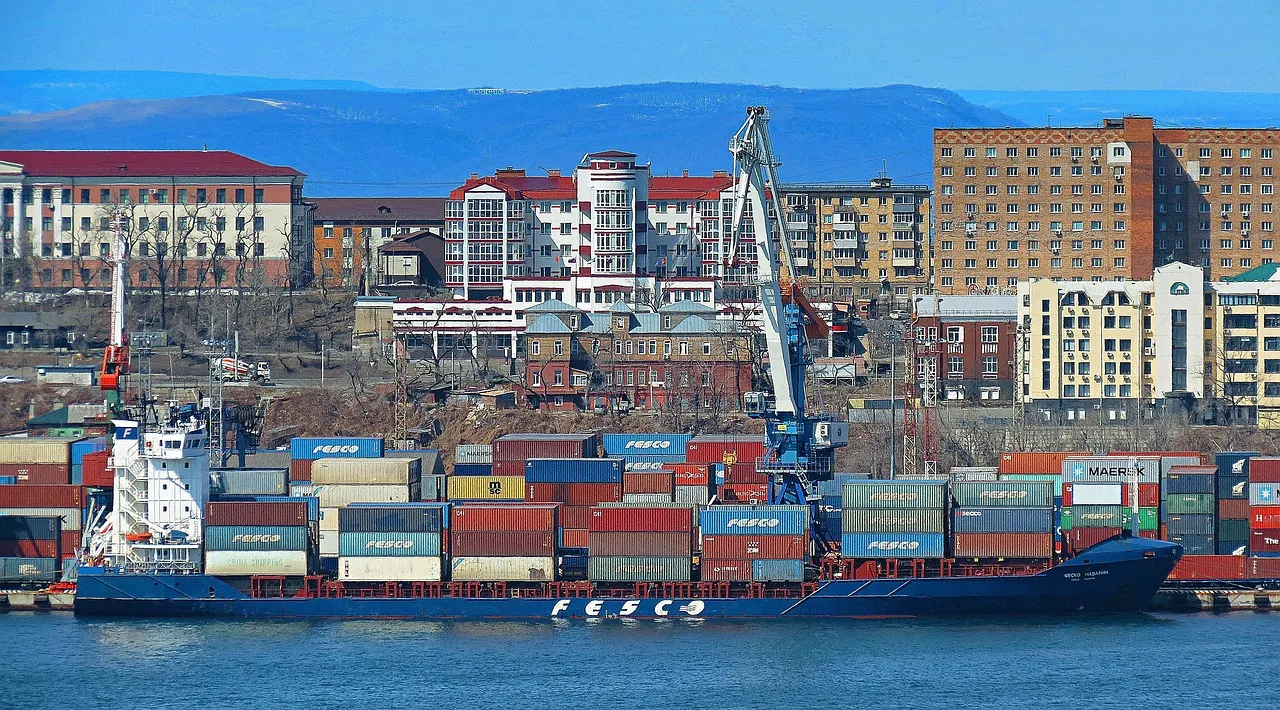 Container ship alongside stacked containers beneath a hillside city, photograph tagged Vladivostok